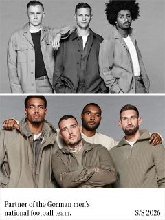 Un hombre con cabello oscuro y rizado, que lleva un blazer negro sobre un jersey de punto verde, está de pie con los brazos cruzados frente a un fondo gris liso y mira al frente con una expresión neutral.