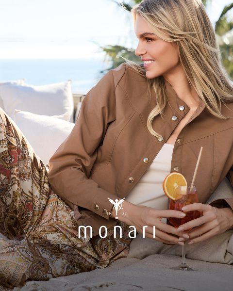 Una mujer sonriente con un abrigo beige, una camisa a rayas, vaqueros azules y una bufanda clara está de pie al aire libre frente a un edificio alto. El logo de Monari se ve en la parte inferior de la imagen.