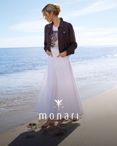 Una mujer sonriente con un abrigo beige, una camisa a rayas, vaqueros azules y una bufanda clara está de pie al aire libre frente a un edificio alto. El logo de Monari se ve en la parte inferior de la imagen.