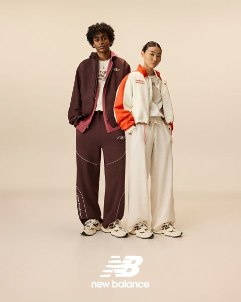 Un hombre y una mujer posan frente a letreros de neón. El hombre lleva un chándal beige y la mujer un mono vaquero. Ambos llevan zapatillas New Balance. La escena está iluminada por varias luces de neón en azul y rojo.