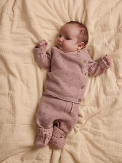 Un bebé está acostado sobre una manta blanca, vistiendo un suéter de punto rosa claro con cuello Peter Pan y pantalones a juego, así como calcetines color crema.