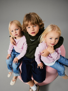 Dos niños pequeños están de pie, tomados de la mano, frente a un fondo marrón liso. Un niño lleva una chaqueta rosa y una falda, el otro jeans azules y una camisa azul con la inscripción "EL AMOR ES NIÑO". Ambos miran hacia arriba con curiosidad.