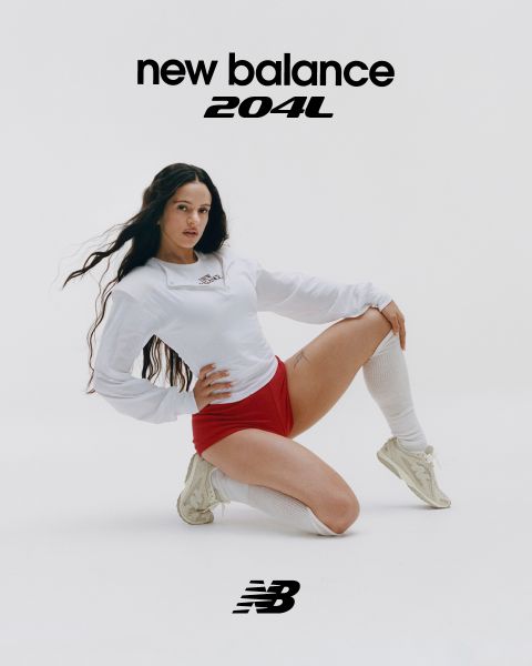 Un hombre y una mujer posan frente a letreros de neón. El hombre lleva un chándal beige y la mujer un mono vaquero. Ambos llevan zapatillas New Balance. La escena está iluminada por varias luces de neón en azul y rojo.