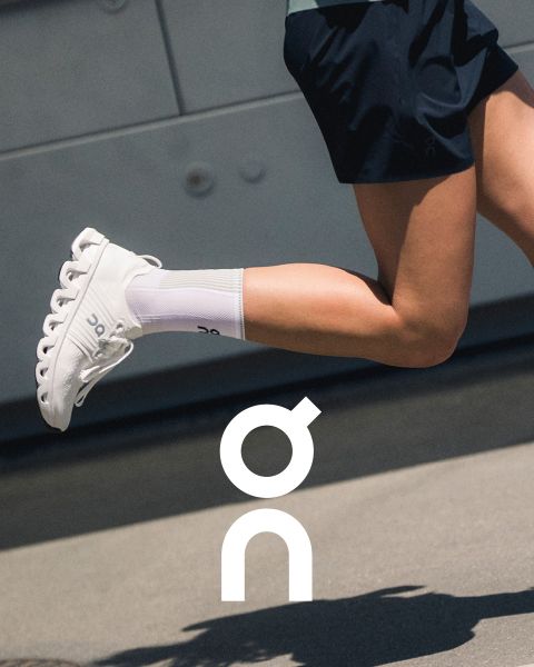 Dos zapatillas de la marca "On" se presentan sobre una rama texturizada frente a un cielo de atardecer colorido. Una zapatilla es negra con detalles grises y la otra es blanca con detalles más oscuros. Debajo, se ve el logotipo de "On".