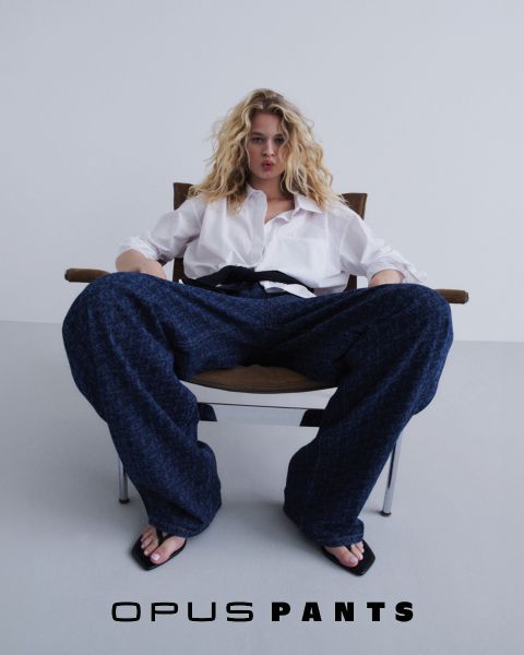 Persona con camisa blanca holgada y vaqueros azules, con las manos en las caderas. Abajo está el texto "Opus Pants". El cabello está ligeramente despeinado por el viento, fondo blanco.