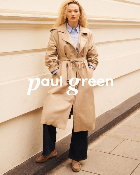 Una persona con pelo largo y rojo, gafas de sol, una blusa verde claro, pantalones beige y zapatillas azules está sentada en un terreno rocoso bajo un cielo azul. Las palabras "Paul Green" están superpuestas en la imagen.