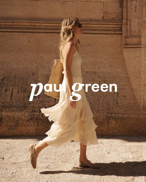 Una persona con pelo largo y rojo, gafas de sol, una blusa verde claro, pantalones beige y zapatillas azules está sentada en un terreno rocoso bajo un cielo azul. Las palabras "Paul Green" están superpuestas en la imagen.