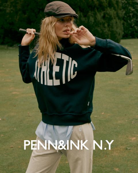 Una mujer con cabello largo y ondulado lleva un traje oscuro y sandalias de tacón alto, y está de pie con confianza en un estudio con un fondo de un solo color. El texto en la parte inferior dice "PENN&INK N.Y."
