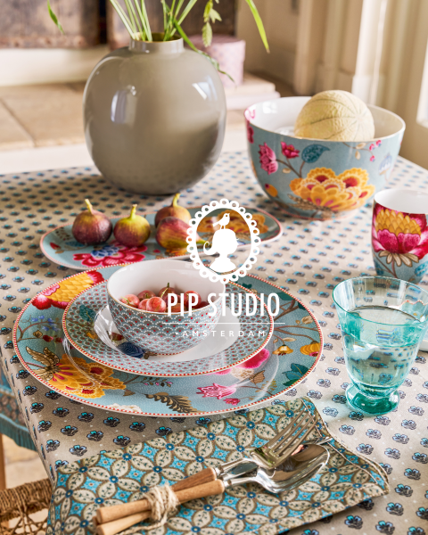 Sobre una mesa con un mantel de rayas verdes hay jarrones de cristal con ramas frondosas y flores rosas. Platos con diseños elaborados se colocan junto a los cubiertos. En el fondo se puede ver una silla de mimbre y un logotipo de Pip Studio.