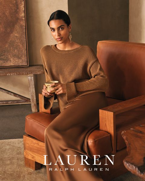 Una mujer con cabello largo y ondulado se reclina en un sofá blanco, vistiendo una blusa roja brillante y pantalones a juego. La cálida luz del sol proyecta sombras sobre la escena. En la parte inferior se lee "LAUREN RALPH LAUREN".