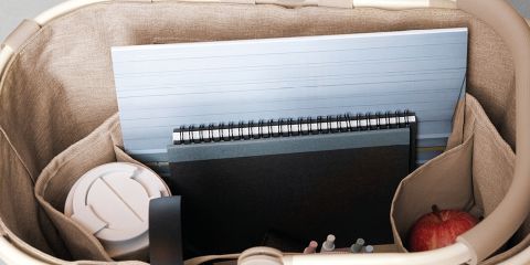 Una persona con cabello largo y rojo, que lleva una camisa azul claro y pantalones blancos, se relaja en el césped y sostiene una bolsa estampada en la mano. Está de pie al aire libre en un campo soleado con vistas a las colinas en la distancia. La palabra "Reisenthel" se ve en letras rojas.