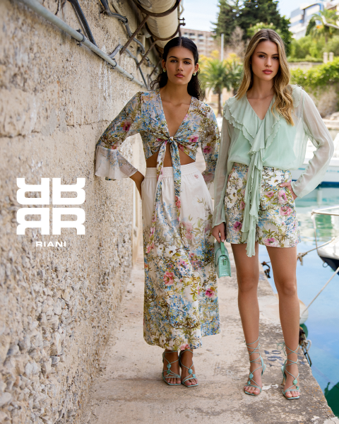 Dos mujeres posan con estilo en un teatro con asientos rojos. Una está sentada en el respaldo de un asiento, vestida con un vestido estampado, un abrigo negro y botas. La otra está de pie junto a ella con un jersey azul marino y una falda negra. Encima de ellas aparece el logo "RIANI".