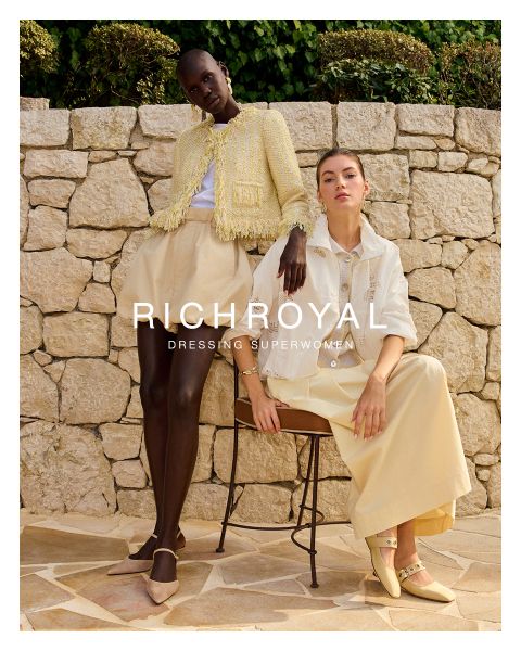 Una mujer con una camisa azul claro y pantalones grises sostiene una bola de nieve frente a un ojo. Posando en una habitación con paredes de madera y un póster de esquí, lleva pendientes grandes. El texto dice "RICHROYAL DRESSING SUPERWOMEN".