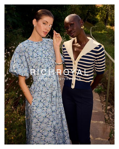 Una mujer con una camisa azul claro y pantalones grises sostiene una bola de nieve frente a un ojo. Posando en una habitación con paredes de madera y un póster de esquí, lleva pendientes grandes. El texto dice "RICHROYAL DRESSING SUPERWOMEN".