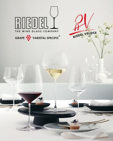Seis coloridas copas de cristal con intrincados patrones tallados están dispuestas sobre una superficie de mármol oscuro. Encima, destaca el logotipo de Riedel con el nombre de la marca y un símbolo de copa de vino.