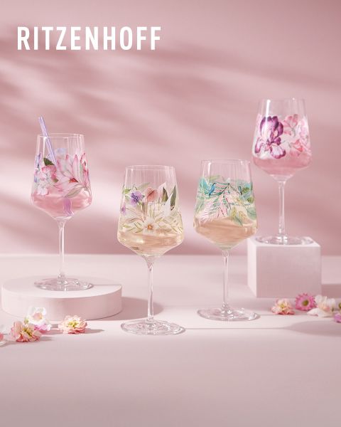 Un conjunto de coloridas y artísticas copas de Ritzenhoff, llenas de bebidas. Cada copa tiene un diseño único con patrones y figuras vibrantes. El fondo muestra reflejos de luz y más copas, creando un ambiente festivo.
