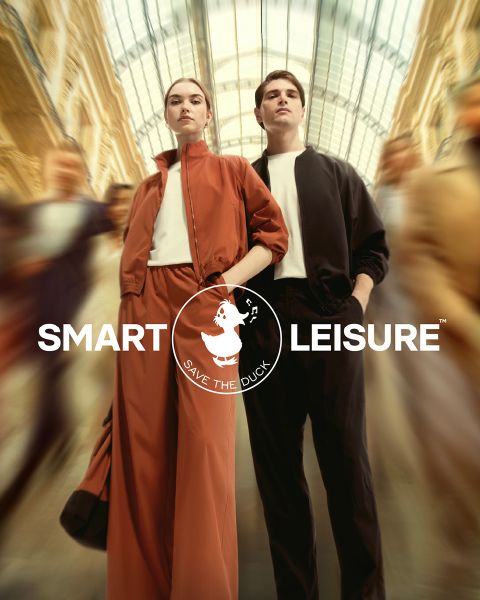 Una modelo camina por un sendero cubierto de hojas. Lleva una chaqueta de piel naranja, una falda a juego y un gorro con medias naranjas y calcetines color melocotón. En la esquina se ve el logo de "Save the Duck".