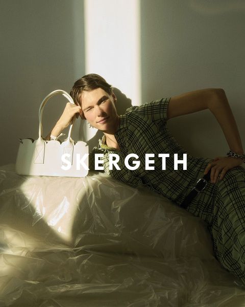 Persona con un blazer negro sobre una camisa a rayas y jeans azules, sosteniendo un bolso negro. El texto "JULIA SKERGETH" es claramente visible superpuesto a la imagen.