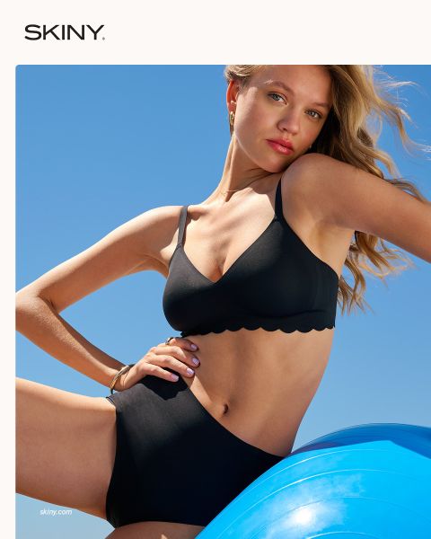 Una mujer con el pelo trenzado se abrocha un sujetador rosa claro con cierre frontal delante de un fondo rojo oscuro. La palabra "SKINY" aparece en la parte inferior de la imagen.