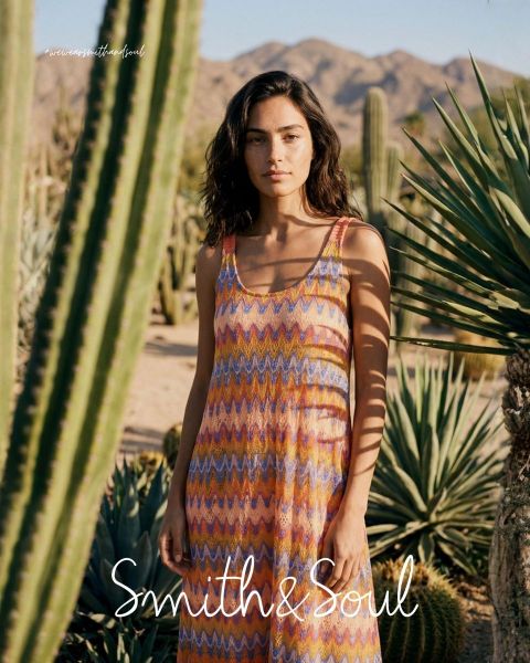 Una mujer con una camisa rosa y una chaqueta de punto con pantalones blancos posa delante de un fondo blanco. El texto dice "Smith & Soul" en la parte inferior.
