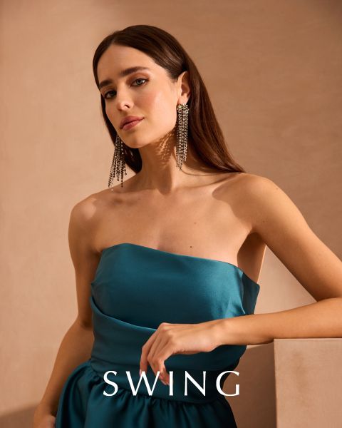 Una mujer con un vestido de seda verde salvia está sentada en una silla color crema frente a un fondo floral. Lleva tacones dorados, pendientes llamativos y una pulsera. La palabra "SWING" aparece en la parte inferior de la imagen.