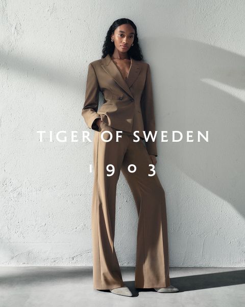 Una mujer con un traje gris está de pie en un muelle de madera en un lago con orillas rocosas y arboladas. El texto "TIGER OF SWEDEN 1903" está escrito a través de la imagen.