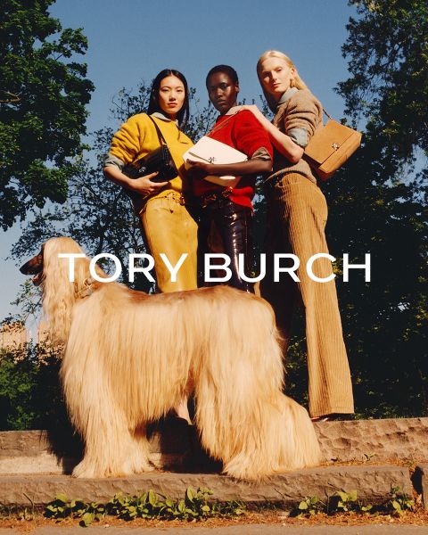 Una mujer con una blusa brillante y una falda verde está sentada junto a cajas de regalo verdes y sostiene un bolso Tory Burch con flores en la mano; detrás de ella hay un árbol de Navidad decorado.