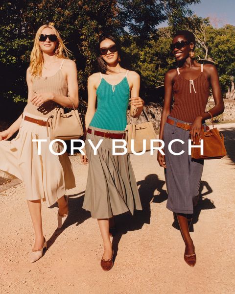 Una mujer con una blusa brillante y una falda verde está sentada junto a cajas de regalo verdes y sostiene un bolso Tory Burch con flores en la mano; detrás de ella hay un árbol de Navidad decorado.