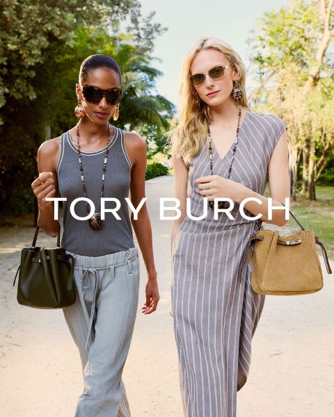 Una mujer con una blusa brillante y una falda verde está sentada junto a cajas de regalo verdes y sostiene un bolso Tory Burch con flores en la mano; detrás de ella hay un árbol de Navidad decorado.