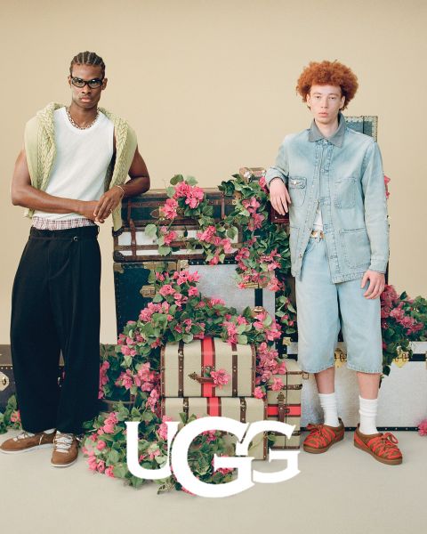 Un par de botas UGG de gamuza marrón con un forro interior de felpa, descansando sobre una superficie gris. El logo de UGG está prominentemente colocado en la parte inferior de la imagen.