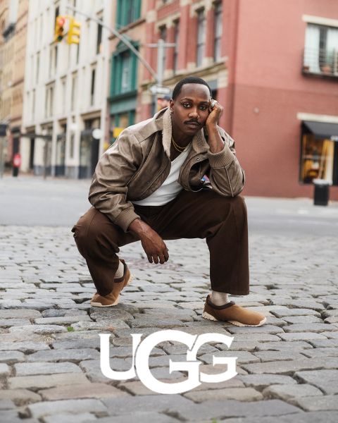 Un par de botas UGG de gamuza marrón con un forro interior de felpa, descansando sobre una superficie gris. El logo de UGG está prominentemente colocado en la parte inferior de la imagen.