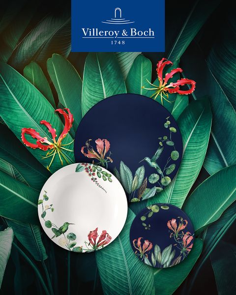 En una mesa de centro de cristal reflectante hay una taza de café en una taza blanca y dorada, una pequeña tetera, una jarrita de leche, una vela y un plato con galletas. La escena parece elegante y acogedora, y en la parte inferior se ve el logotipo de Villeroy & Boch.