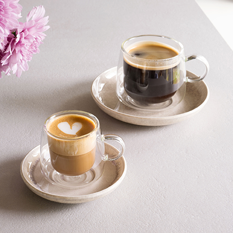 Un vaso de vidrio de doble pared está lleno de yogur cremoso y adornado con frambuesas, moras y arándanos frescos. El vaso está sobre un plato negro con una cuchara al lado, delante de un fondo oscuro.