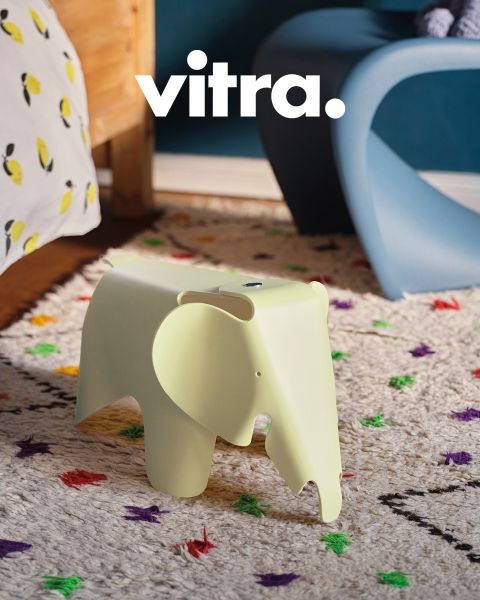 Un salón moderno cuenta con un sofá a rayas rosa y beige, una silla blanca con patas de madera y una mesa de centro redonda de madera. Sobre la mesa hay un jarrón con flores coloridas, encima pone "vitra." en la pared. El suelo está cubierto con una alfombra suave.