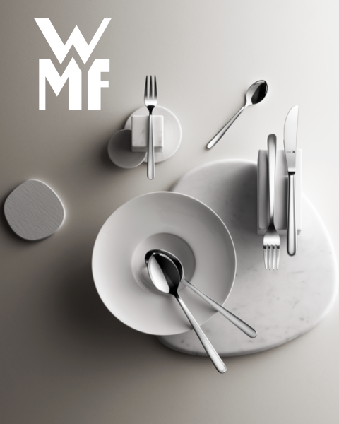 Una olla llena de higos, moras y guarniciones aromáticas está sobre una tabla de madera oscura. Encima de la olla se ve el logo de WMF. El fondo es una superficie oscura texturizada.