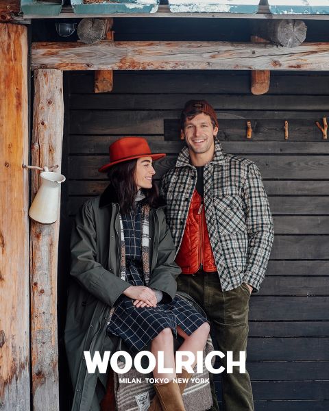 Una persona con un abrigo de invierno marrón claro, un gorro de punto azul y una camisa de cuadros azules sobre una sudadera con capucha está de pie frente a un fondo liso. Debajo aparece la palabra "Woolrich" junto con las ciudades de Milán, Tokio y Nueva York.