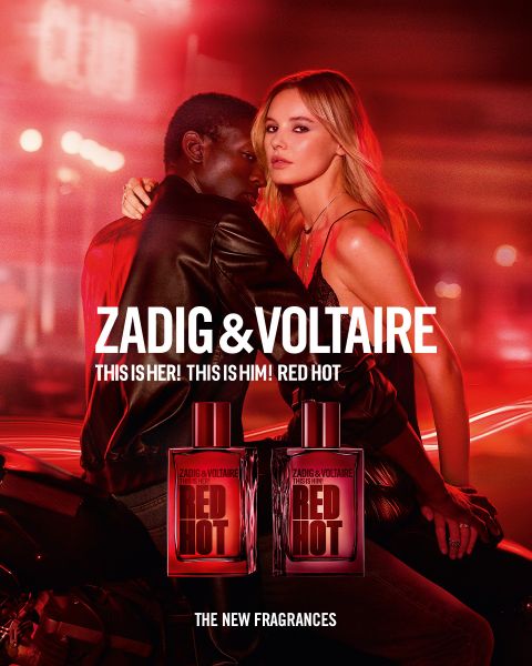 Mujer con cabello largo y rojo y un atuendo negro sentada en una cama en una habitación con una jaula de pájaros y una ventana grande. Texto: "Zadig & Voltaire" y "Zadig: La nueva fragancia recargable". Logotipo con alas y una herradura.