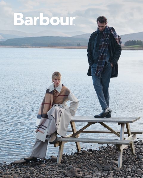 Dos personas con elegante ropa Barbour de varias capas y bufandas a cuadros posan junto a un lago. Uno está sentado en una mesa de picnic, mientras que el otro está de pie, con montañas y agua al fondo. El logotipo de Barbour aparece en la esquina superior izquierda.
