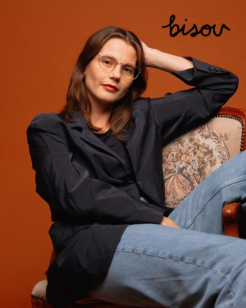 Una mujer con gafas, blazer oscuro y vaqueros está sentada en una silla ornamentada frente a un fondo color óxido, encima de ella está la palabra "Bisou" en letras negras.