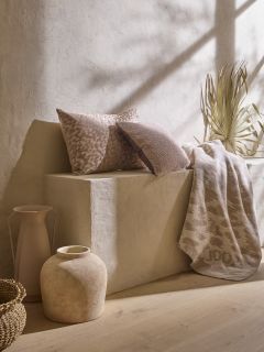 Un espacio minimalista presenta un banco de piedra beige con dos cojines marrones y beige texturizados, una manta estampada y hierbas secas. Una cesta trenzada, un jarrón de cerámica rústica y una jarra alta adornan el suelo bajo una suave luz natural.