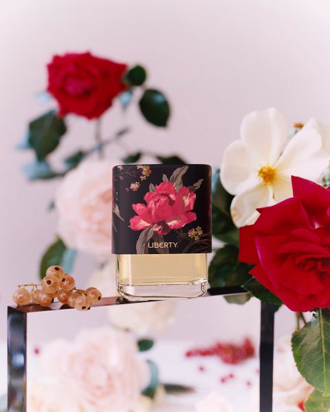 Frasco de perfume con diseño floral y la inscripción "Liberty" sobre una superficie de vidrio. Rodeado de rosas rojas y blancas, hojas verdes, grosellas rojas y un fondo borroso con más flores.