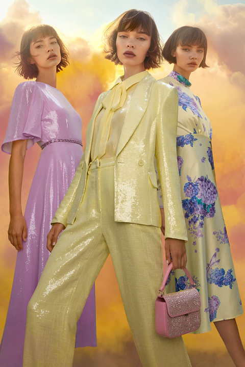 Tres modelos posan frente a un cielo nublado y brillante. Llevan atuendos de color pastel: un vestido lila, un traje de lentejuelas amarillo con pajarita y un vestido blanco de flores. Una modelo sostiene un bolso de mano rosa con brillo.