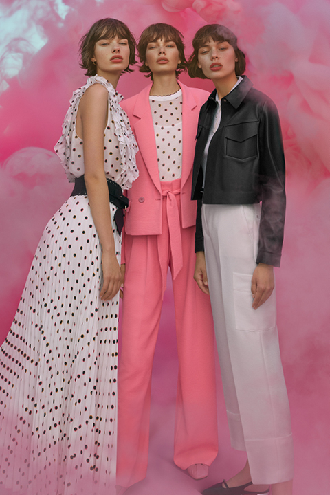 Tres mujeres vestidas a la moda están de pie frente a un fondo rosado y ahumado. Una lleva un vestido de lunares, otra un traje rosa con una camisa de lunares y la tercera una chaqueta negra y pantalones blancos. Posan con confianza y emanan un aire moderno y elegante.
