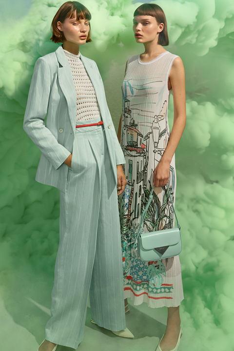 Dos modelos posan frente a un fondo de humo verde. Una lleva un traje azul claro con una blusa estampada en blanco, la otra un vestido sin mangas con estampados gráficos. Ambas tienen el pelo corto y oscuro y llevan accesorios a juego en tonos pastel.