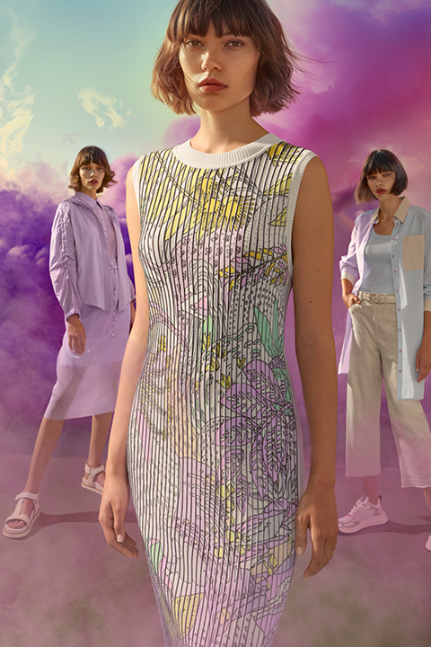 Tres mujeres posan frente a un brillante fondo de nubes púrpuras y rosas. La figura central lleva un vestido estampado, mientras que las otras visten atuendos casuales y de colores claros. La escena es colorida y artística.
