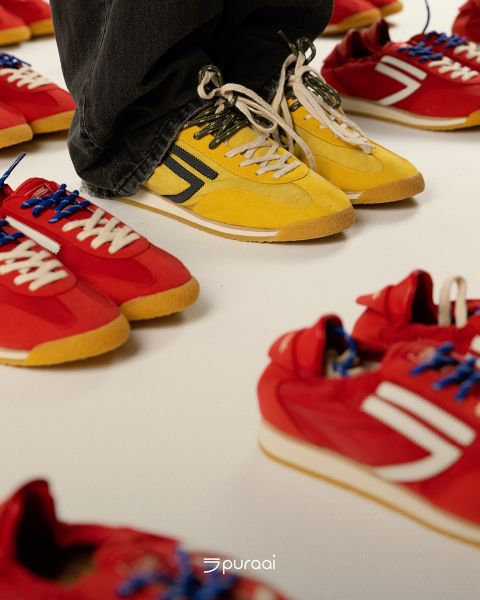 Un par de pies con zapatillas amarillas con cordones blancos se encuentra rodeado de varios pares de zapatillas rojas con cordones azules sobre una superficie blanca.