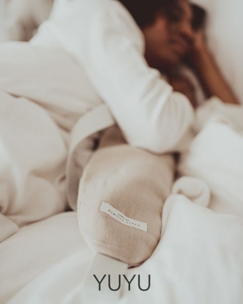 Una persona está acostada en una cama, envuelta en una manta blanca, y sostiene una almohada larga y brillante o una almohadilla térmica en su mano. La atmósfera es tranquila y acogedora, transmitiendo seguridad y relajación. Abajo está la palabra "YUYU".