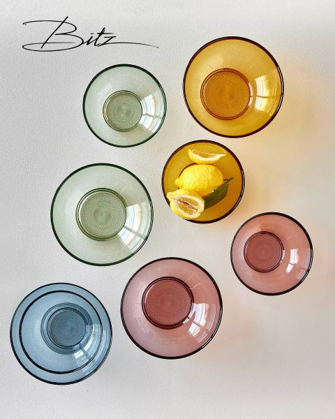 Siete cuencos de vidrio coloridos, dispuestos en círculo sobre una superficie blanca. Un cuenco amarillo contiene tres limones con hojas verdes. El logo de "Bitz" se ve en la esquina superior izquierda.