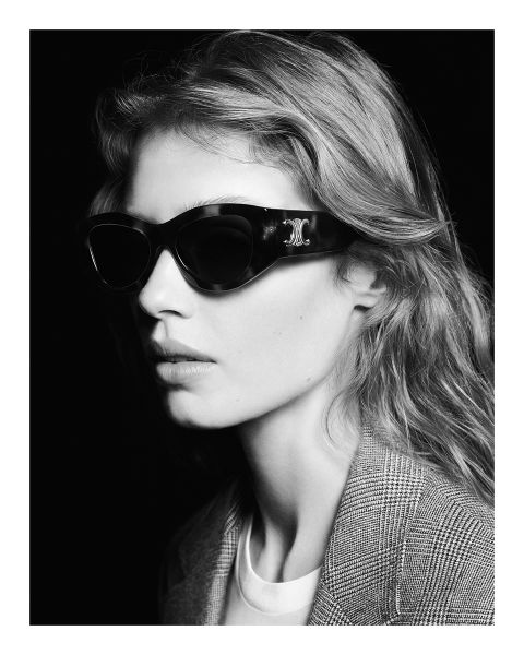 Retrato en blanco y negro de una persona con cabello largo, gafas de sol oscuras y un blazer a cuadros, mirando hacia un lado. La imagen tiene una estética elegante y sofisticada.