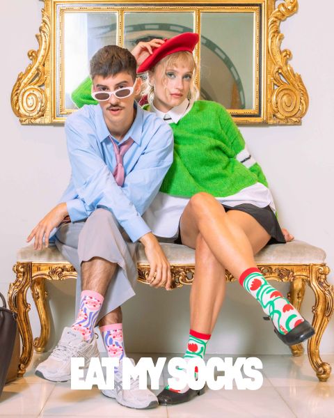 Un joven y una joven están sentados en un banco adornado. Ambos llevan calcetines extravagantes con diseños coloridos. El hombre lleva gafas de sol y una camisa azul, mientras que la mujer lleva una boina roja y un jersey verde. Abajo se puede ver la frase "EAT MY SOCKS".